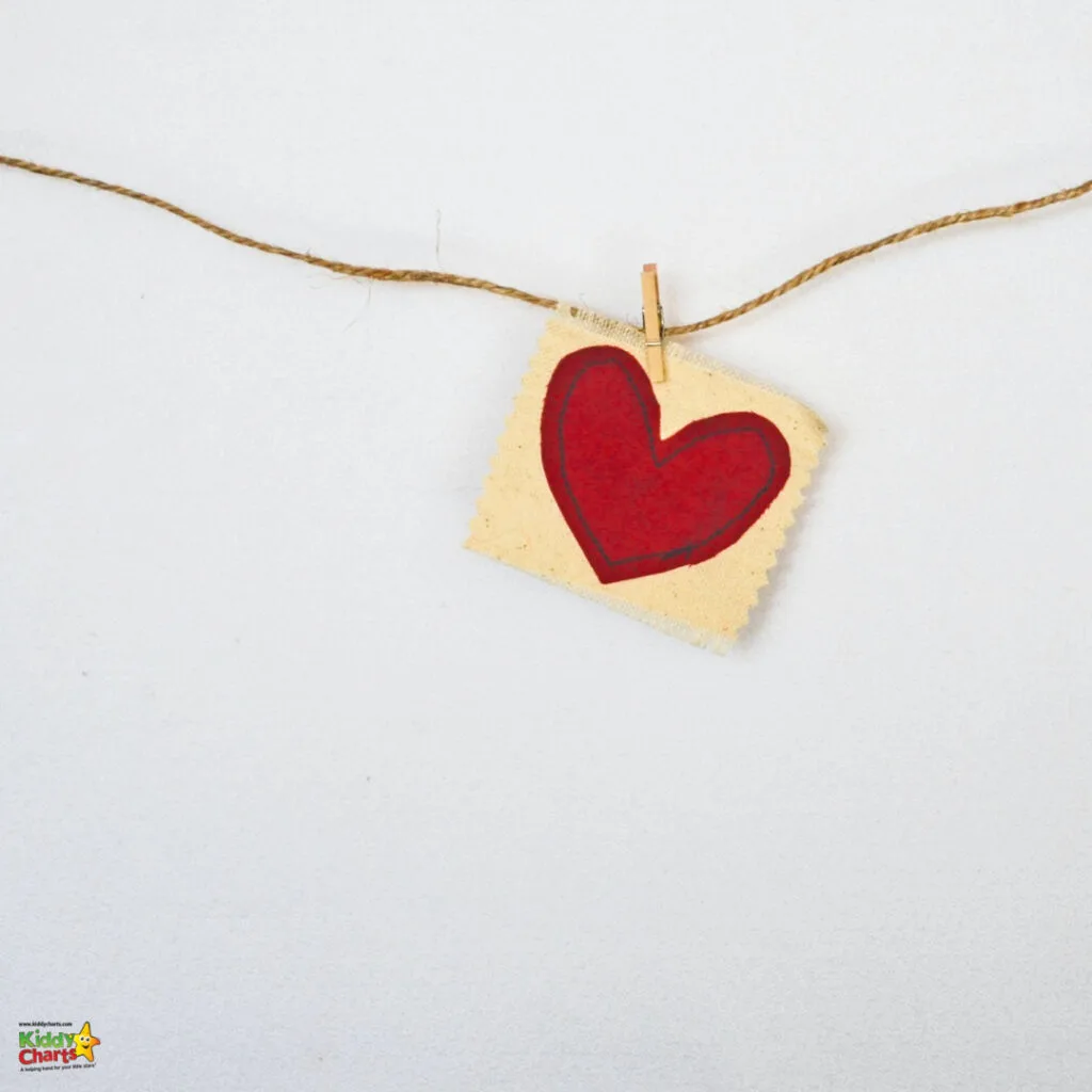 A small square piece of beige fabric with a red heart design is pinned by a wooden clothespin to a twine against a white background.