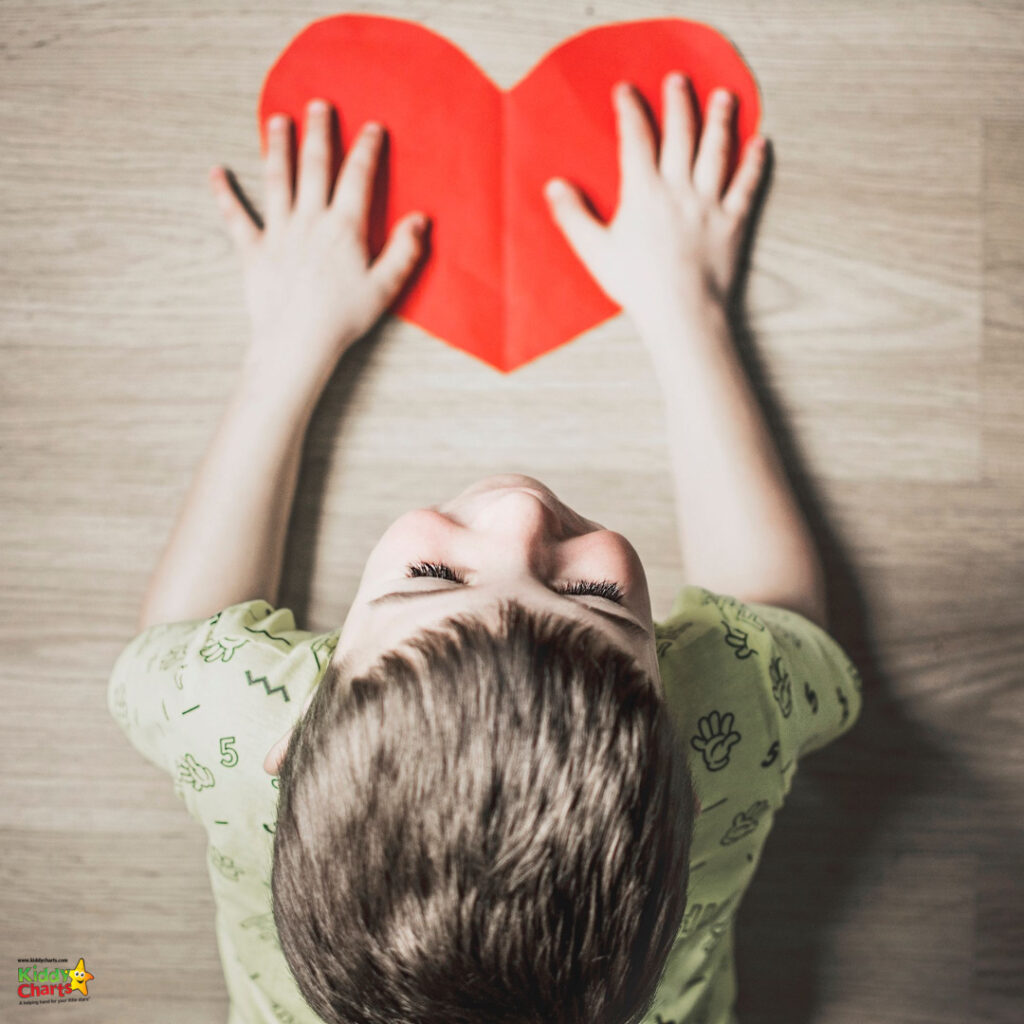 A child lies on a wooden floor, gazing upwards, with hands gently touching a large, folded red paper heart. The atmosphere is calm and contemplative.