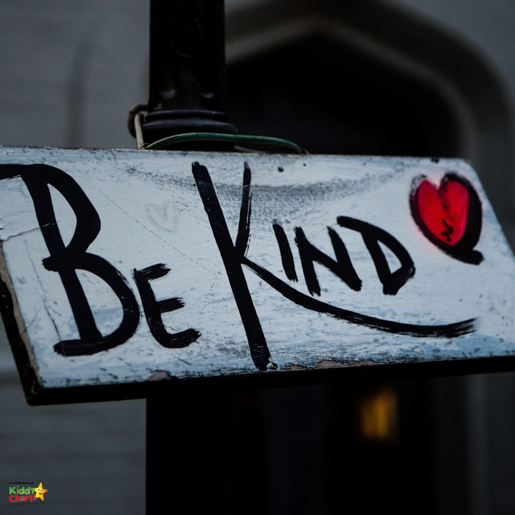 A weathered sign hanging from a lamp post with the handwritten message "Be Kind," accompanied by a small red heart drawing.