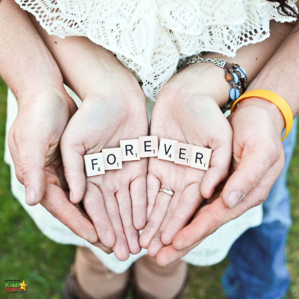 Two people hold hands, showing "FOREVER" spelled with letter tiles. Both wear rings, suggesting a romantic connection, standing on grassy ground.