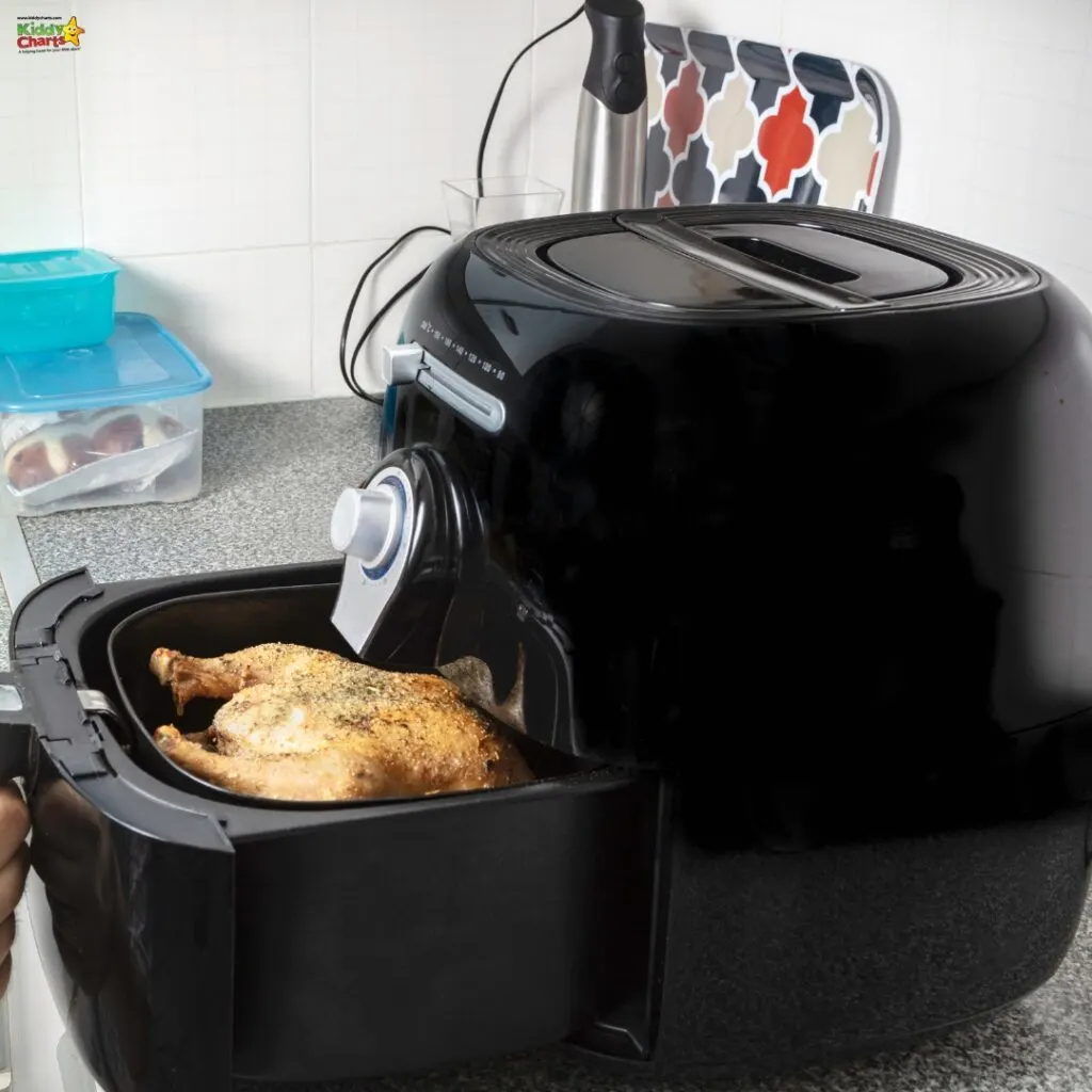 A counter-top air fryer with a whole roasted chicken inside. Kitchen background with a patterned tile wall and food containers.