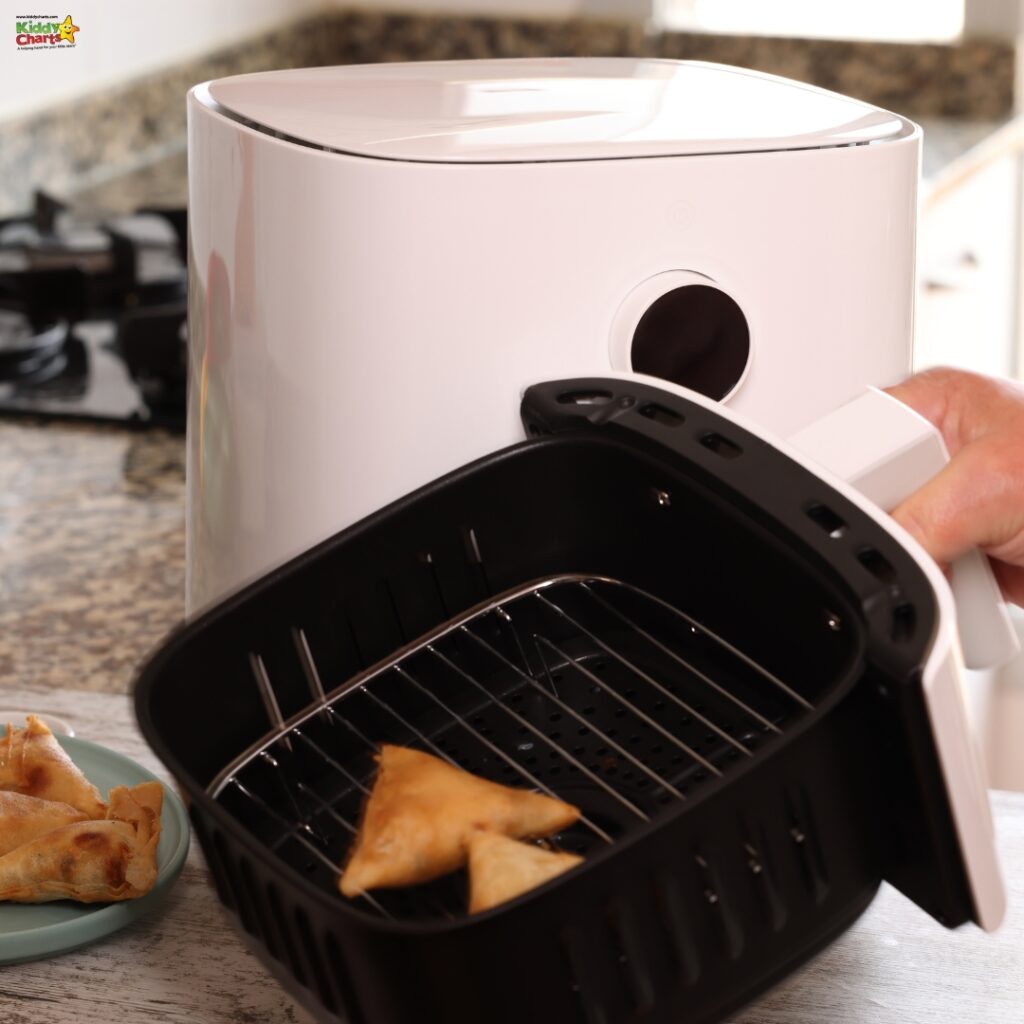 A white air fryer sits on a kitchen counter. A person is pulling out the basket containing golden-brown samosas. A gas stove is in the background.
