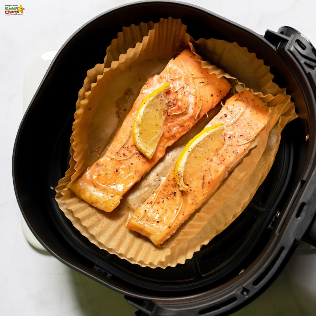 Two seasoned salmon fillets with lemon slices atop are in an air fryer, prepared for cooking on a brown parchment liner. It's a home-cooked meal setup.