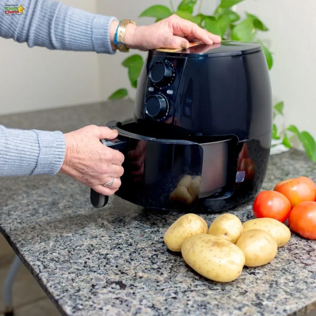 A person uses a black air fryer on a kitchen countertop, surrounded by raw potatoes and tomatoes, indicating meal preparation.