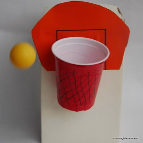 This image shows a homemade basketball hoop made from a red cup with drawn net lines, mounted on a white and orange paper backboard, with a yellow ball mid-air.