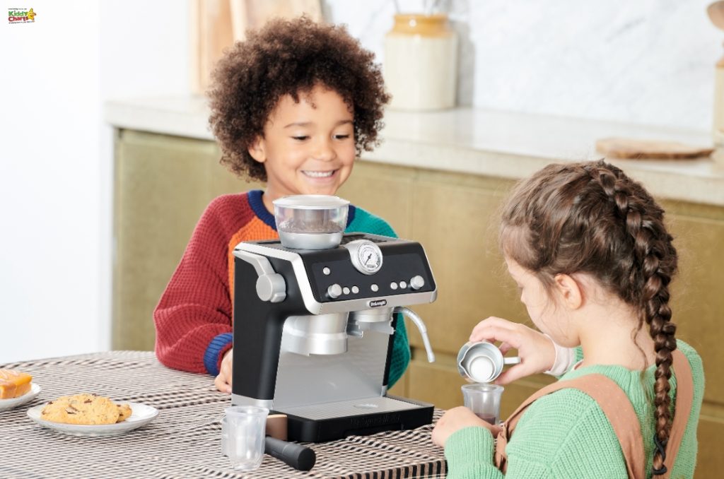 Two children are happily engaging with a toy espresso machine on a kitchen counter. There are snacks nearby, and the setting looks cozy.