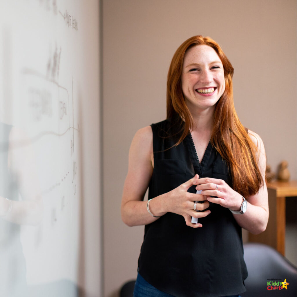 A smiling person with long red hair, wearing a black sleeveless top, stands in front of a whiteboard with charts, exuding confidence and happiness.