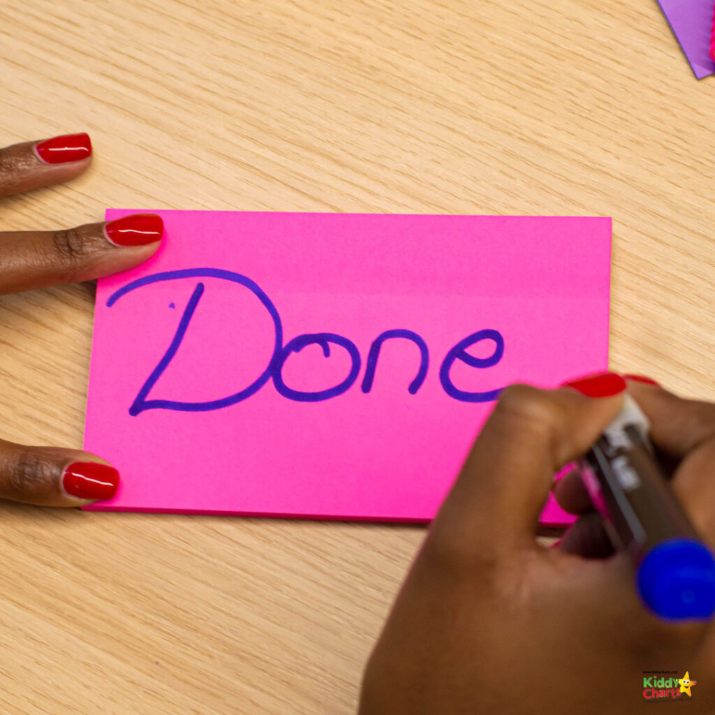 A person is writing the word "Done" on a pink sticky note. Their nails are painted red, and they're holding a blue marker.