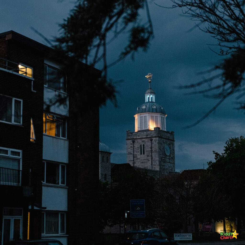 A dusky scene with an illuminated clock tower rising behind an urban building, framed by silhouetted tree branches against a moody evening sky.