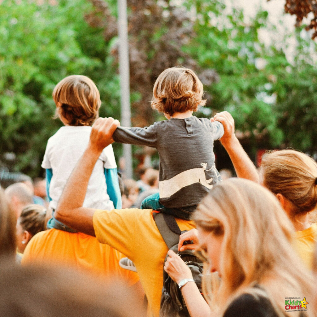 Two children sit atop adults' shoulders in a crowded area, watching an event, surrounded by other attendees, in a warm, sunny outdoor setting.