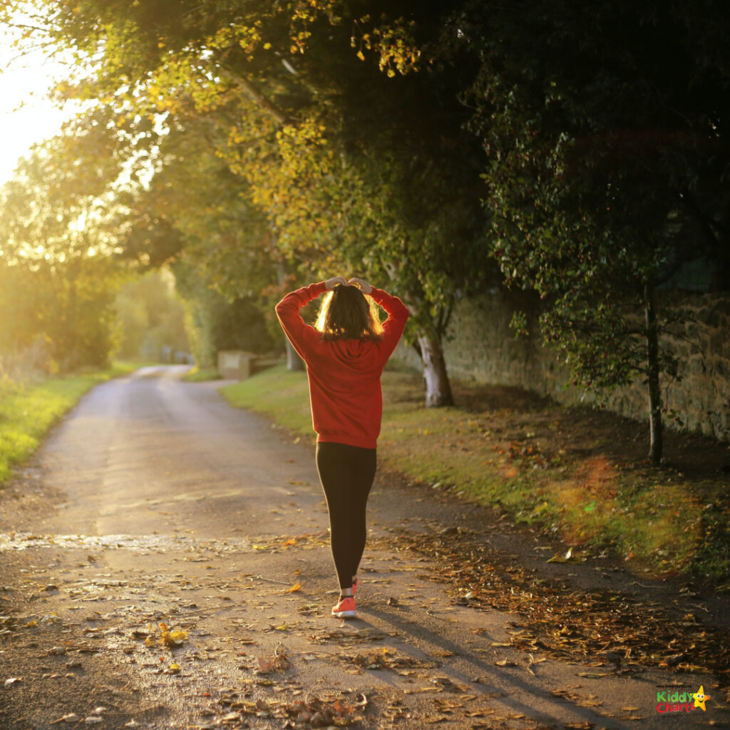 A person is walking along a tree-lined path during sunset, wearing a red hoodie, with hands resting on their head. Golden light filters through leaves.