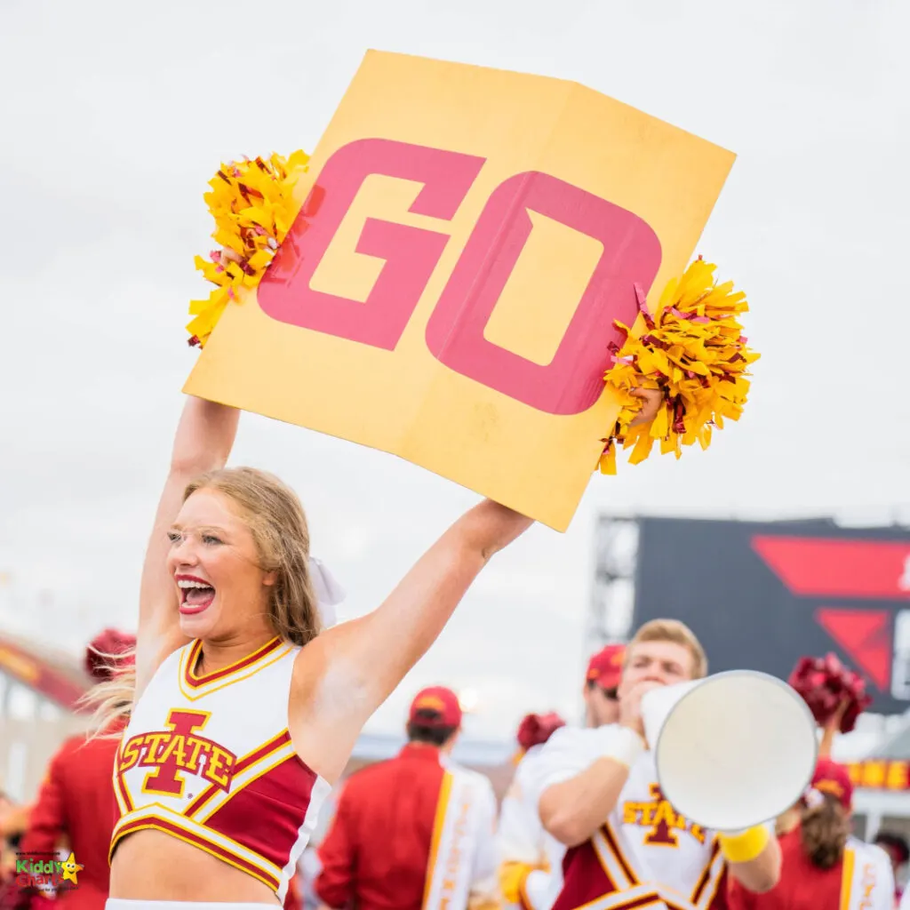 A person in a cheerleader uniform enthusiastically holds up a "GO" sign and pom-poms at an outdoor event, with others in similar attire in the background.