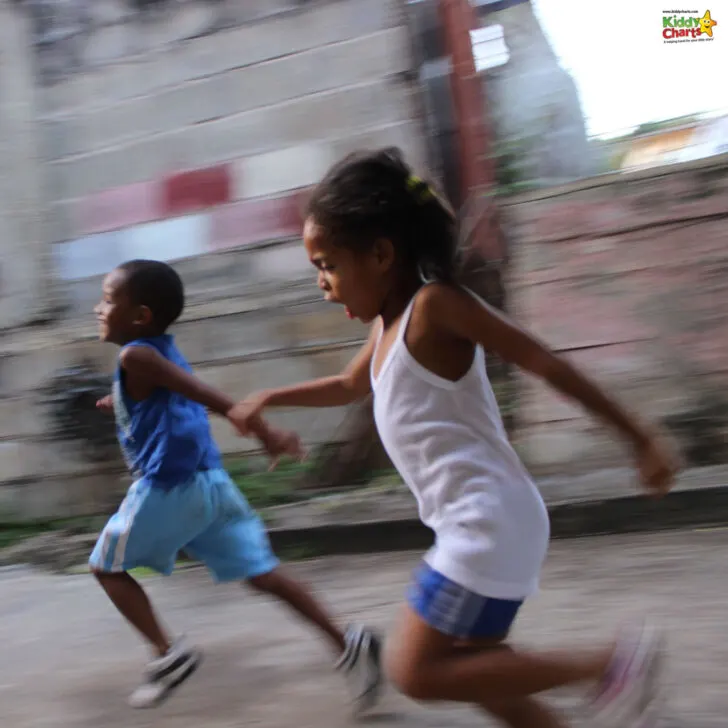 Two children are running joyfully, hand in hand, with a blurred urban background suggesting movement. Both are casually dressed in summer clothing.