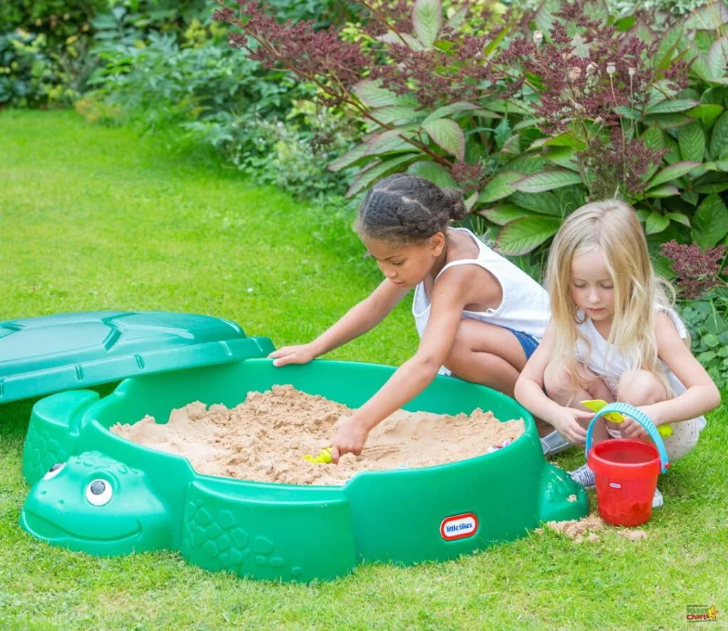 Two children are playing with sand in a green, turtle-shaped sandbox in a lush garden. One child is scooping sand, the other holds a bucket.