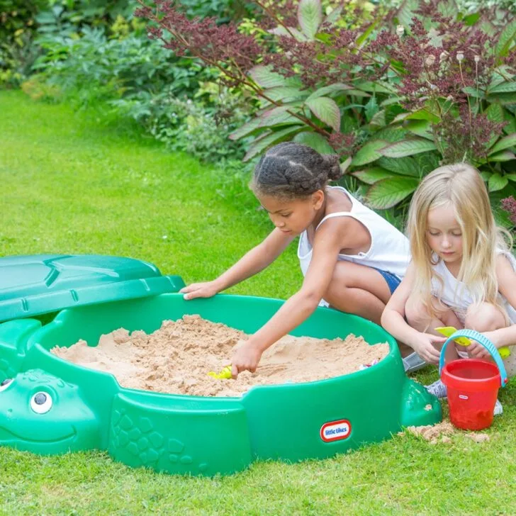 Two children are playing with sand in a green, turtle-shaped sandbox in a lush garden. One child is scooping sand, the other holds a bucket.