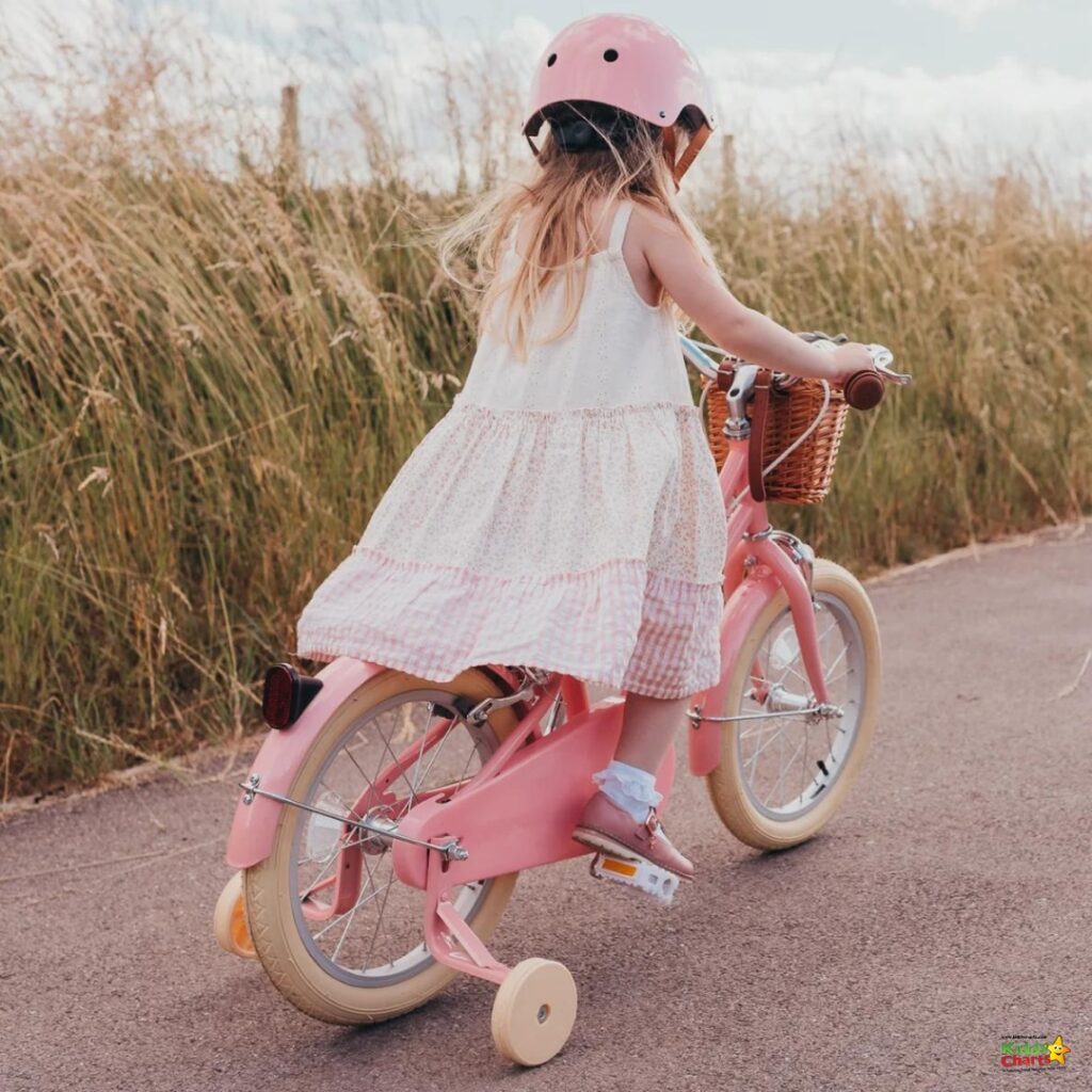 A child in a pink helmet and dress rides a pink bicycle with training wheels on a path by tall grasses, suggesting a leisurely day outdoors.