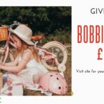 A child sits beside a pink bike, holding flowers, next to a picnic basket and pink helmet, with "GIVEAWAY BOBBIN BIKES £199" text overlay.