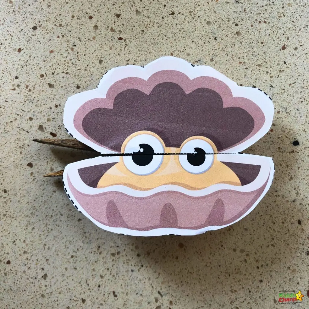 This is a colorful paper cut-out of a clam with a friendly face, big eyes, and a smile, resting on a speckled beige surface.