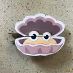 The image shows a whimsical paper cut-out of a cartoon clam with big eyes and a smiling face, resting on a speckled surface.