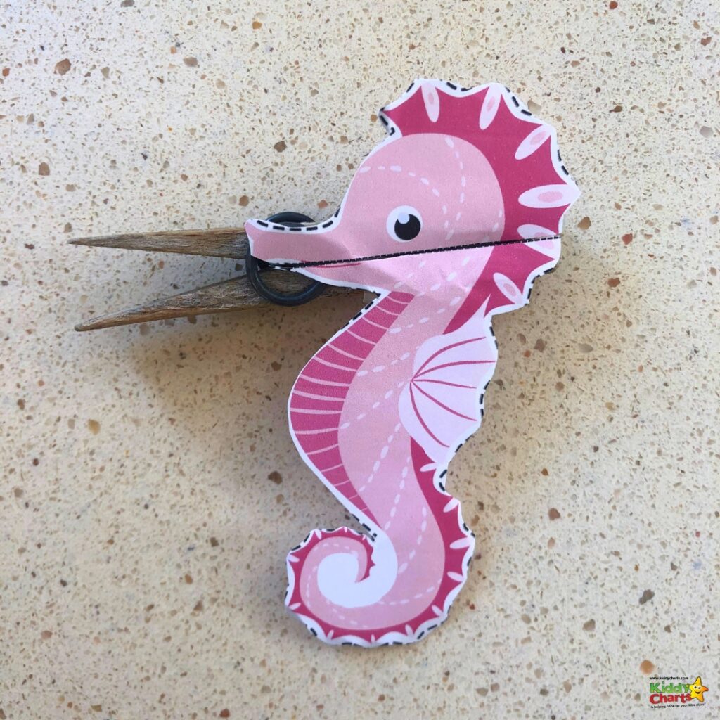 The image shows a pink and white illustrated seahorse clothespin craft, mounted on a textured surface, designed for creative and educational purposes for children.