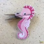 A paper seahorse clip is attached to a clothespin. It's pink with stripes and patterns, creating a playful and decorative look against a speckled background.