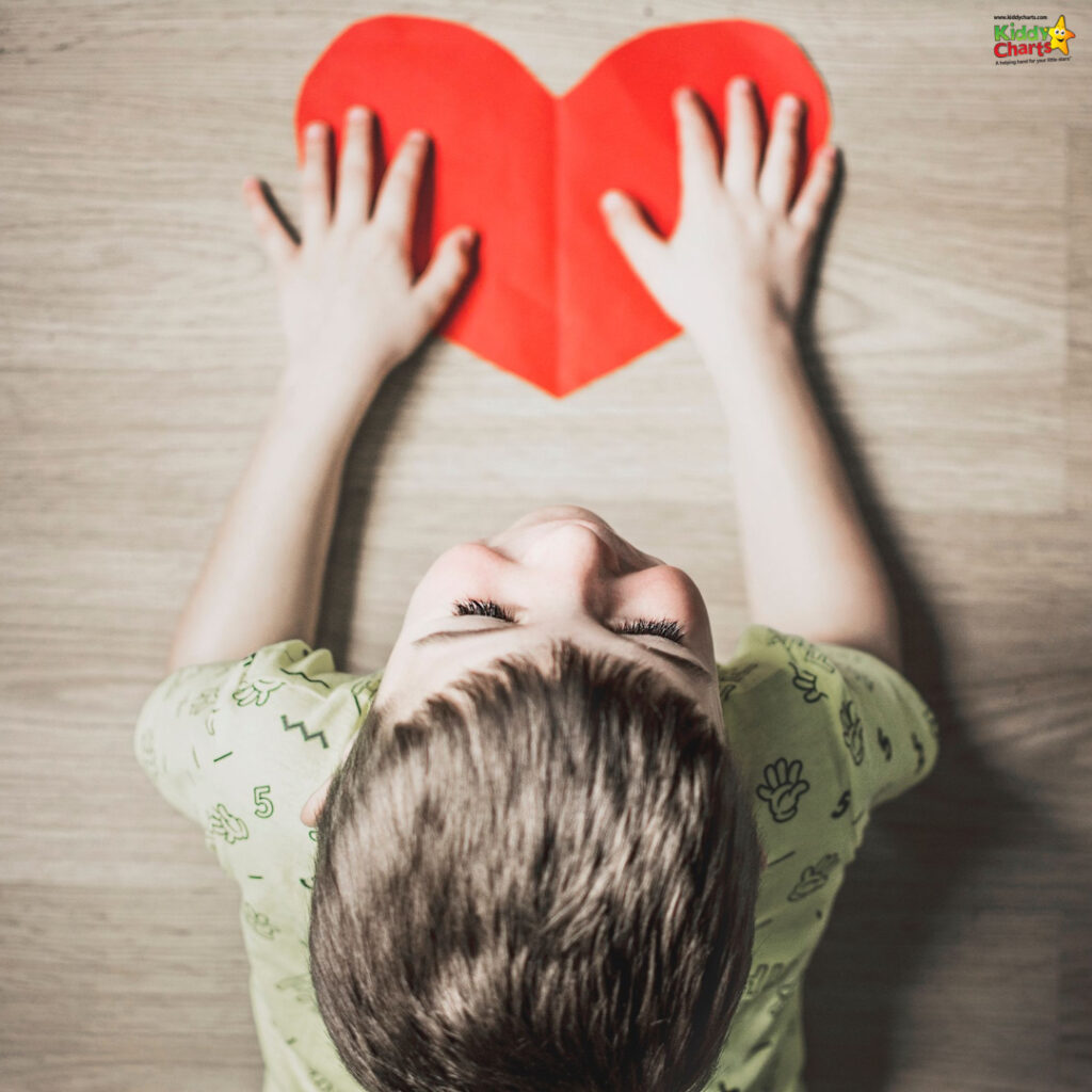 A child lies on the floor, gazing upward with hands extended towards a large red heart shape, suggesting innocence, love, or a playful moment.
