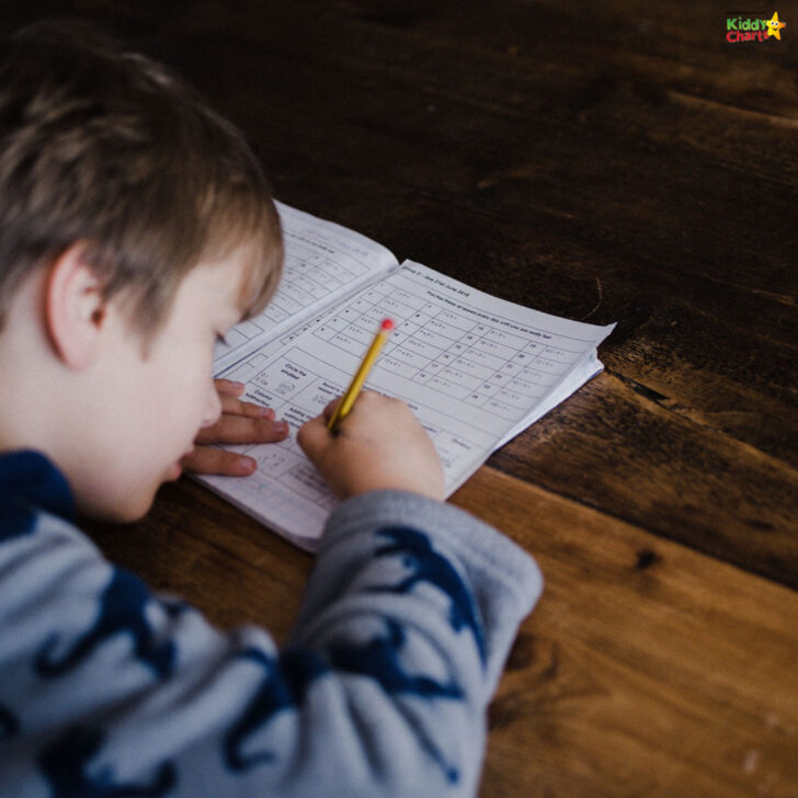 A child in a shark-patterned sweatshirt is focused on writing in a workbook with a yellow pencil on a dark wooden surface.