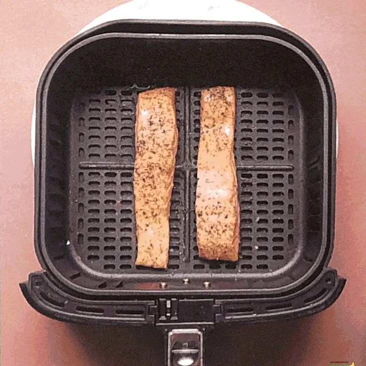 Two slices of bread with visible grill marks are inside an open sandwich maker with a textured surface. The kitchen appliance is against a pink wall.