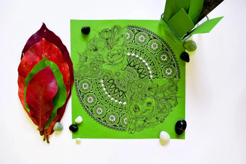 A detailed black mandala design on green paper, surrounded by red and green leaves, and black and white stones.