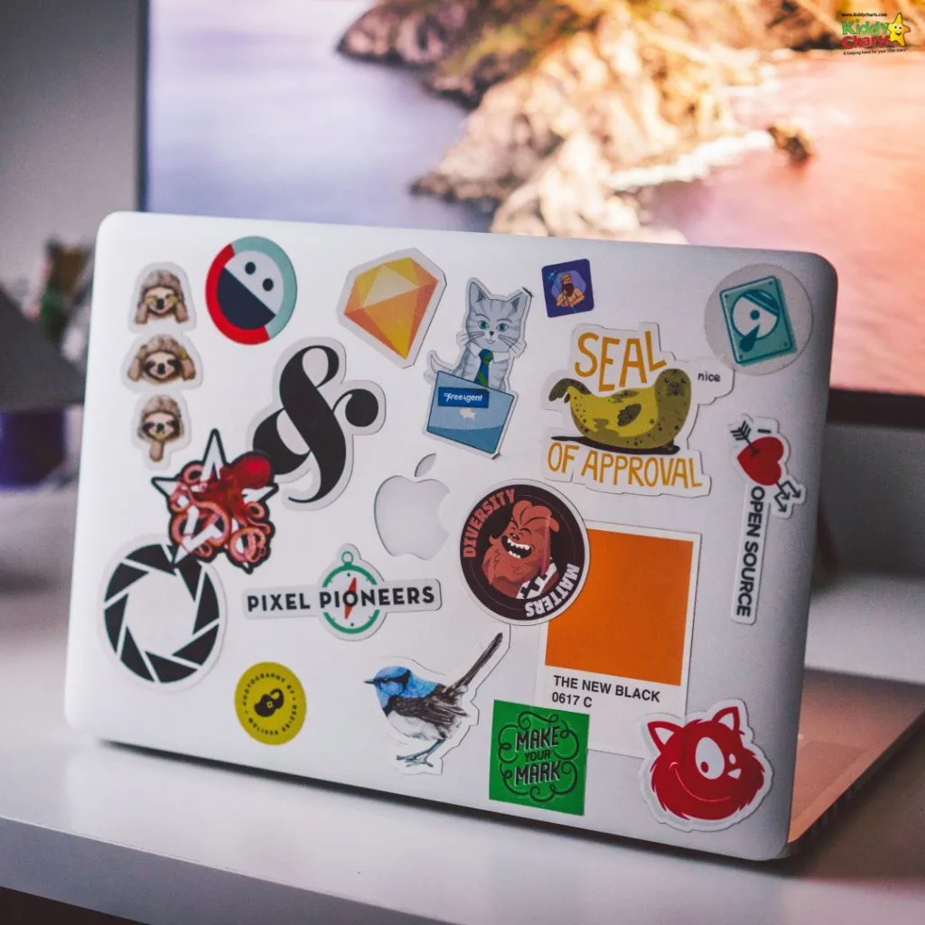 A laptop with various colorful stickers, including seals, cats, and tech logos, sits open on a desk with a blurred background.