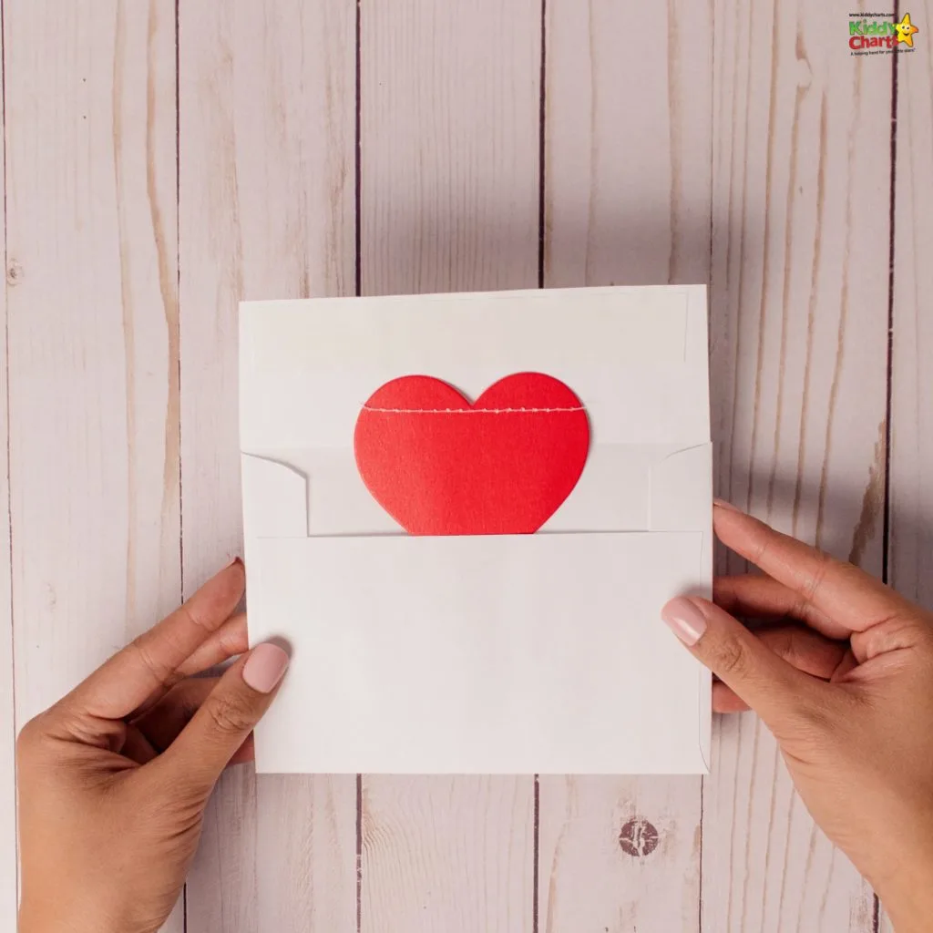 A person holds a white envelope with a red heart card inside, against a wooden background. An emblem is visible in the corner.