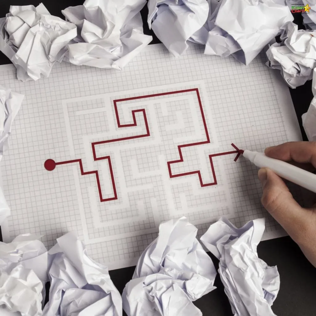 A person draws a path through a maze on graph paper, surrounded by crumpled papers, symbolizing creativity and problem-solving.