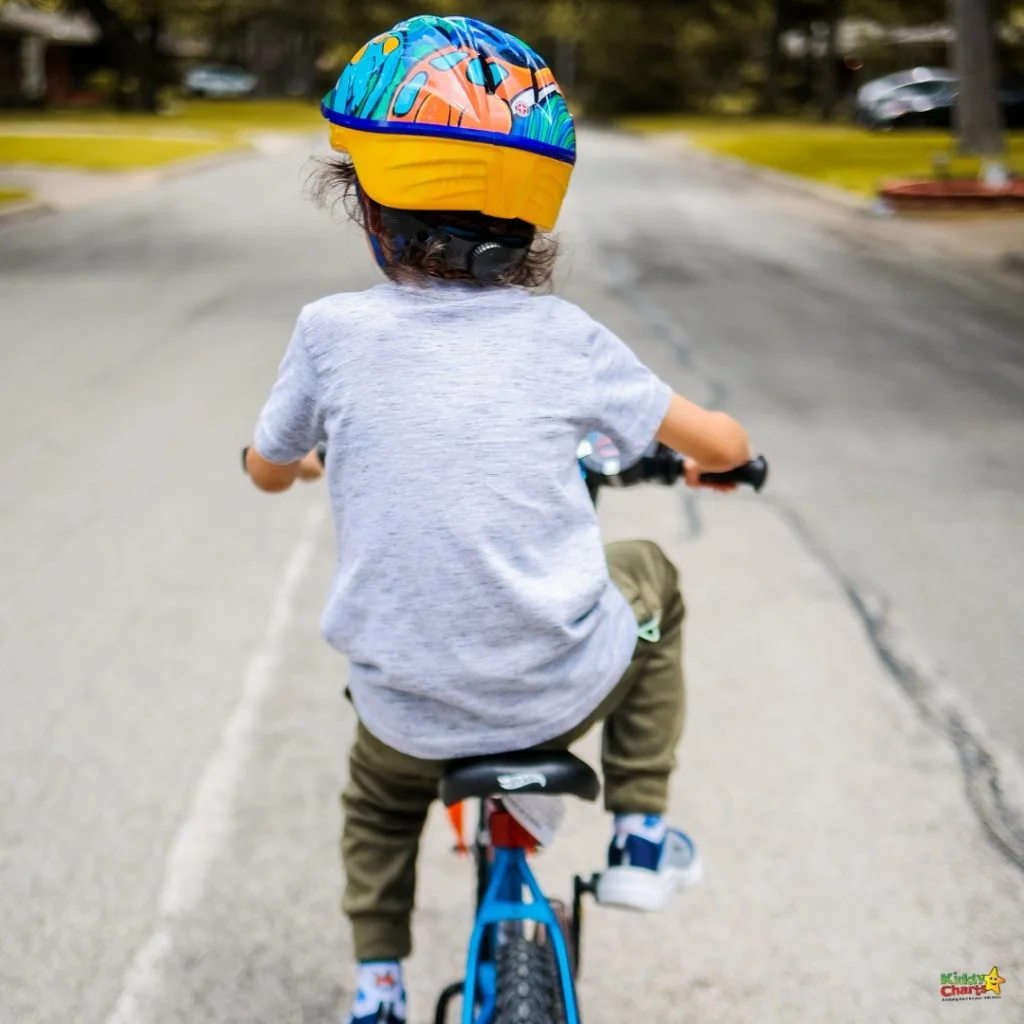 Young child with a colorful helmet rides a bike down a quiet residential street, surrounded by greenery and houses.