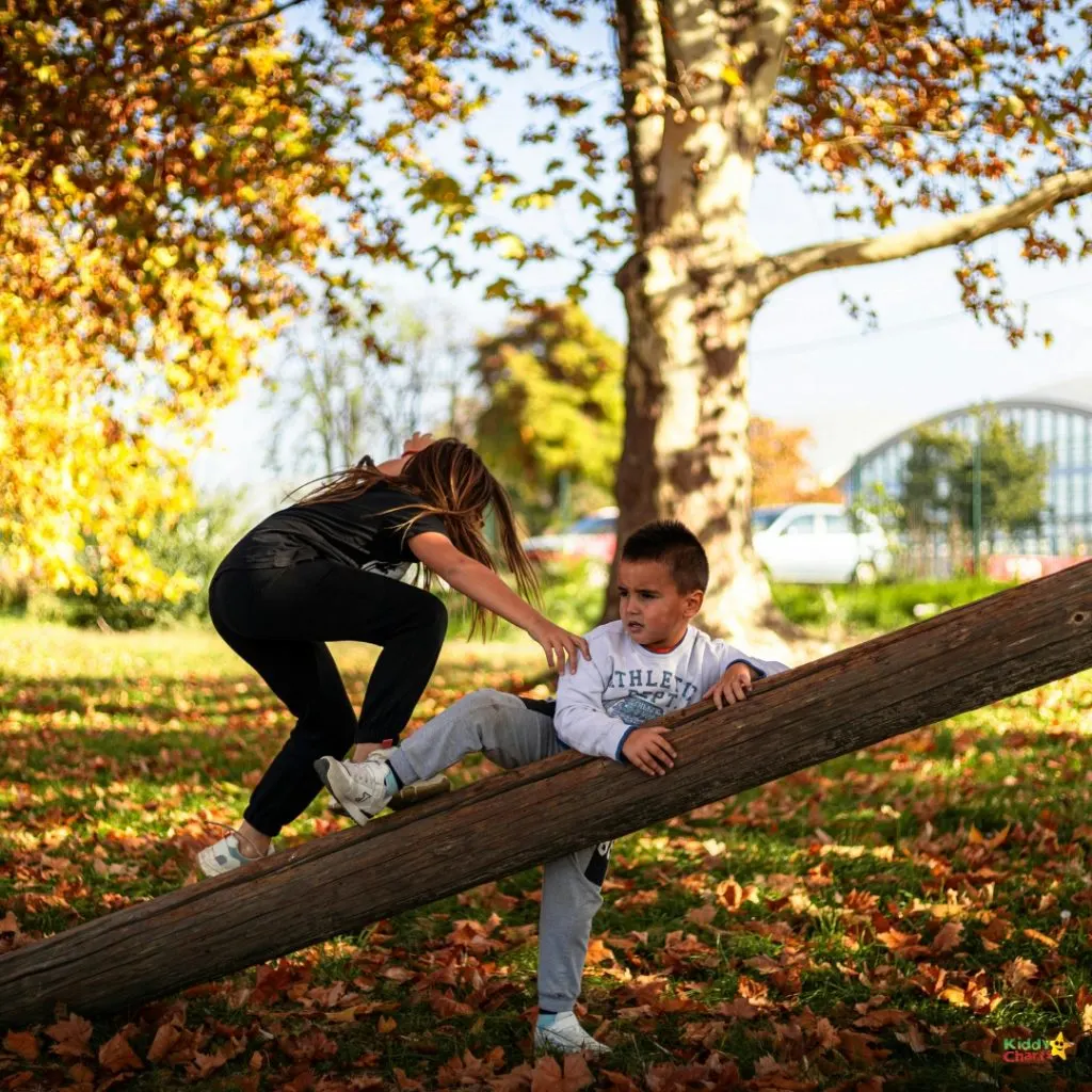 Two children play on a log in an autumn park filled with vibrant leaves, under a large tree, with a building in the background.
