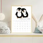 A framed February 2026 calendar featuring two cartoon penguins. Displayed on a shelf with a decorative cloud pillow and rabbit doll.