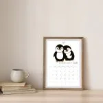 Framed calendar on a table, featuring two cartoon penguins for February 2026. Nearby, a stack of books and a mug sit.
