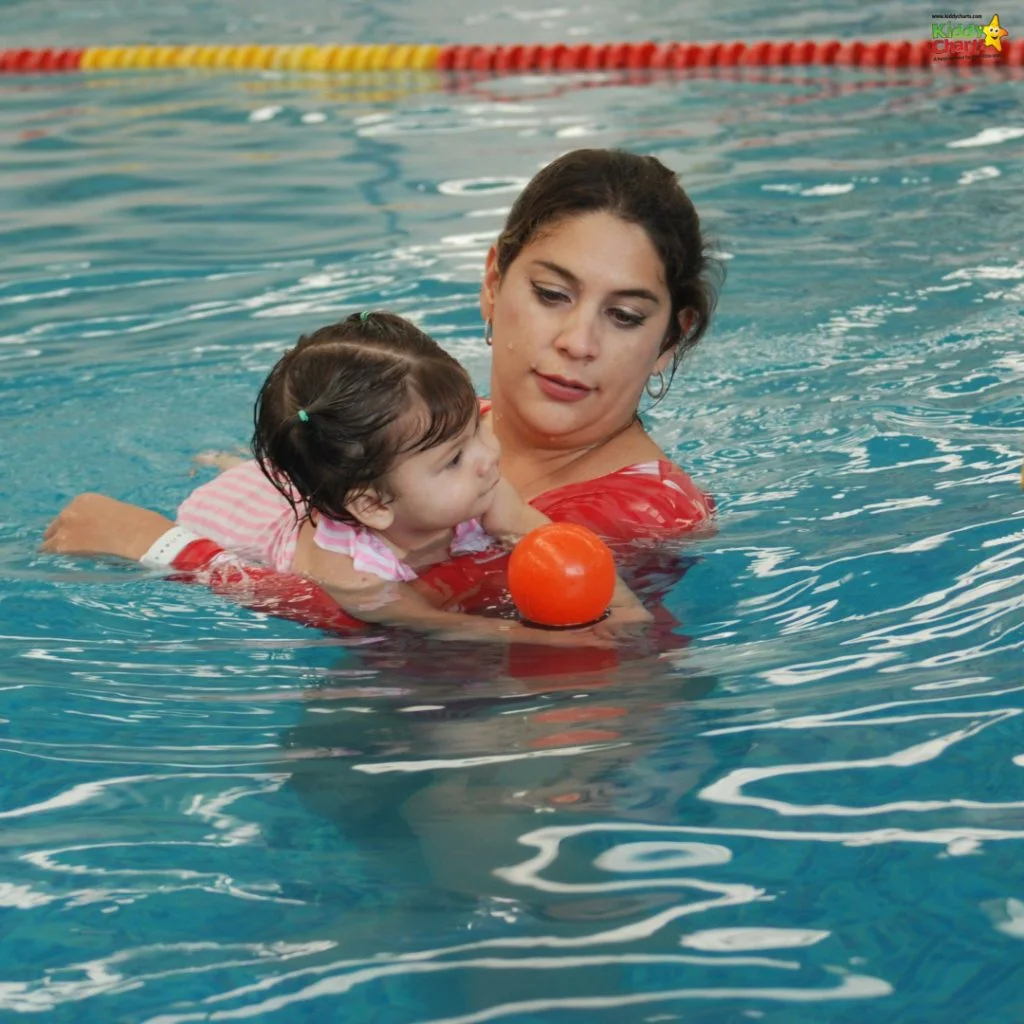 A person helps a child float in a swimming pool, reaching for an orange ball, with lane dividers in the background.