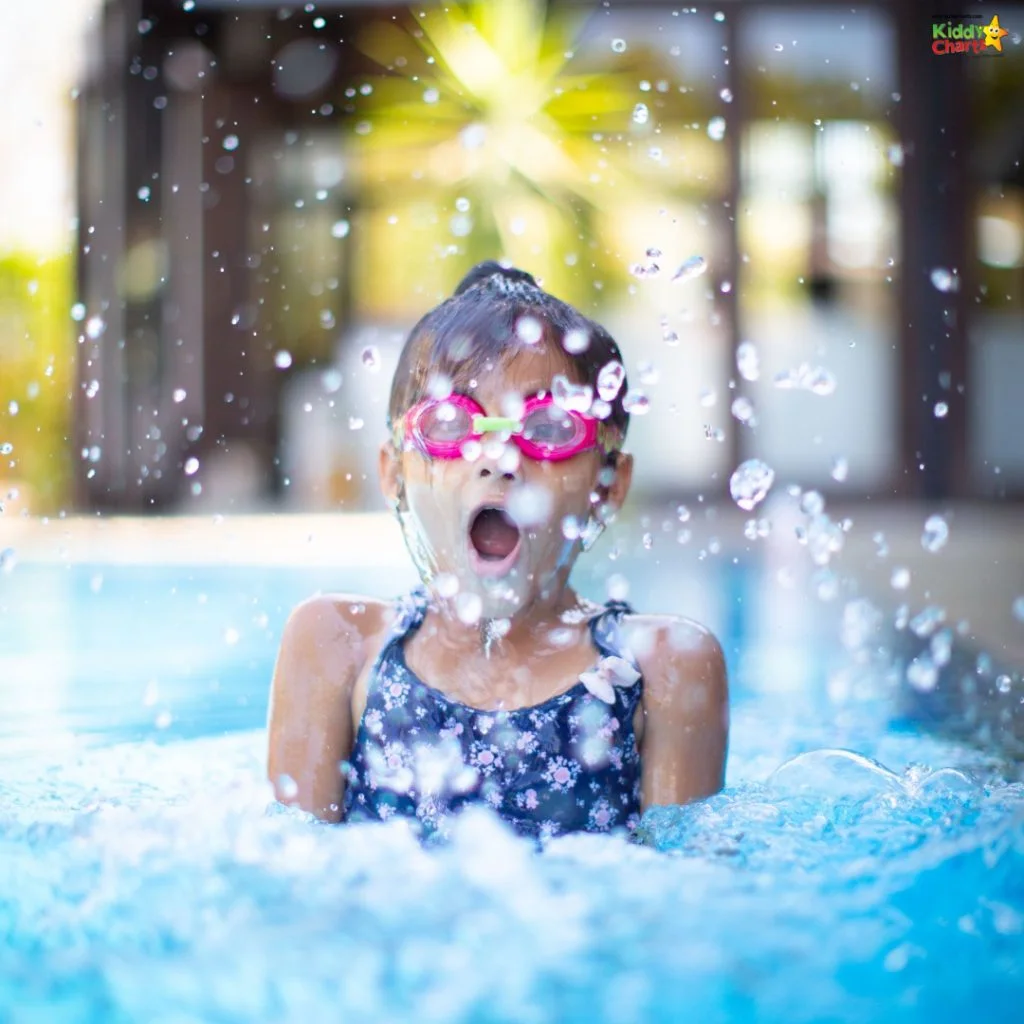 Child wearing pink goggles joyfully splashes in a clear blue pool. Background features blurred greenery and structures, conveying a vibrant, playful atmosphere.