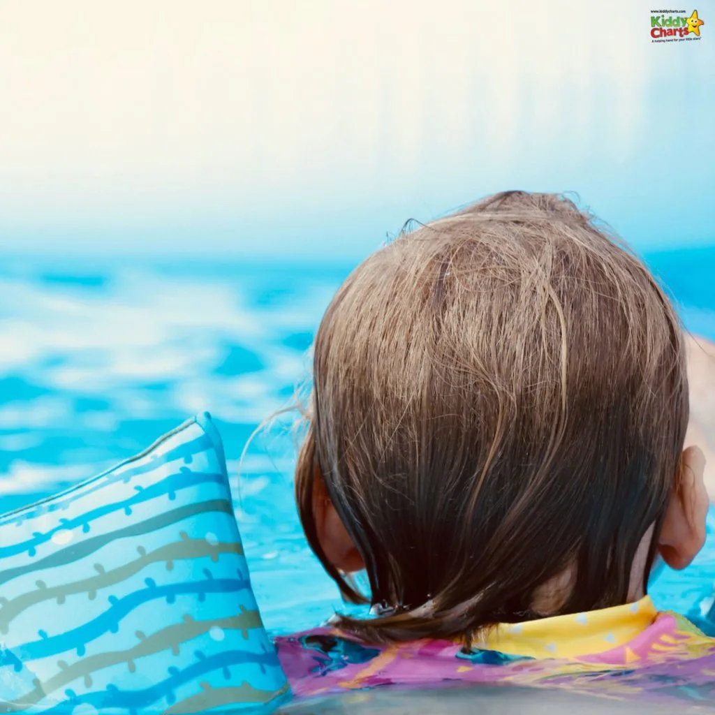 A child wearing a colorful floaty is swimming in a pool with a blurred background and bright blue water.