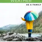 A person in a yellow raincoat holds a colorful umbrella by a river, surrounded by misty, forested mountains.