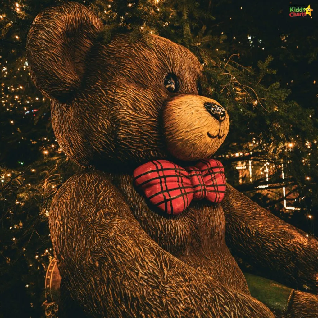 A large teddy bear sculpture with a red plaid bowtie sits near twinkling lights, surrounded by greenery, creating a festive atmosphere.