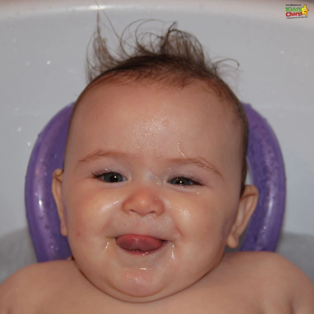 A happy baby with wet hair sits in a bath, smiling gleefully, surrounded by a white and purple bath seat.