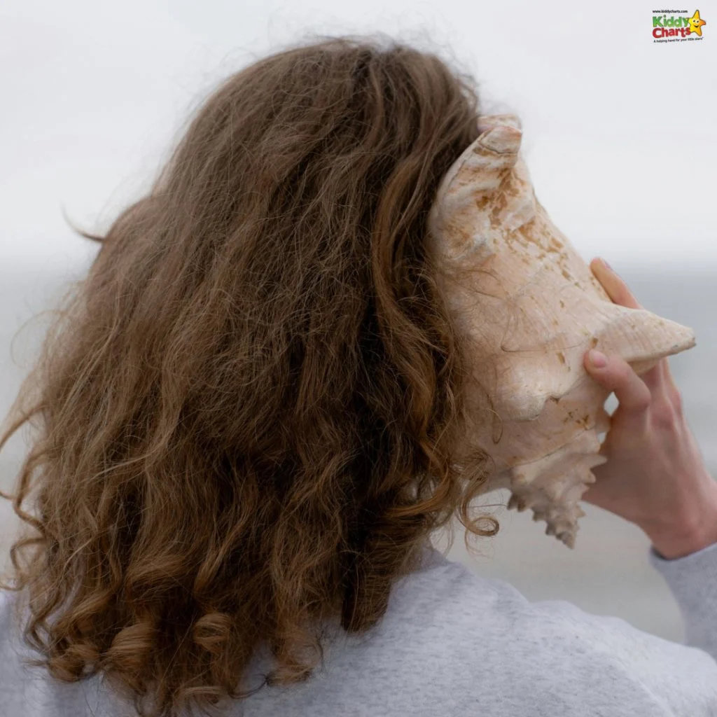 A person with curly hair holds a large conch shell to their ear, standing near the ocean under an overcast sky.