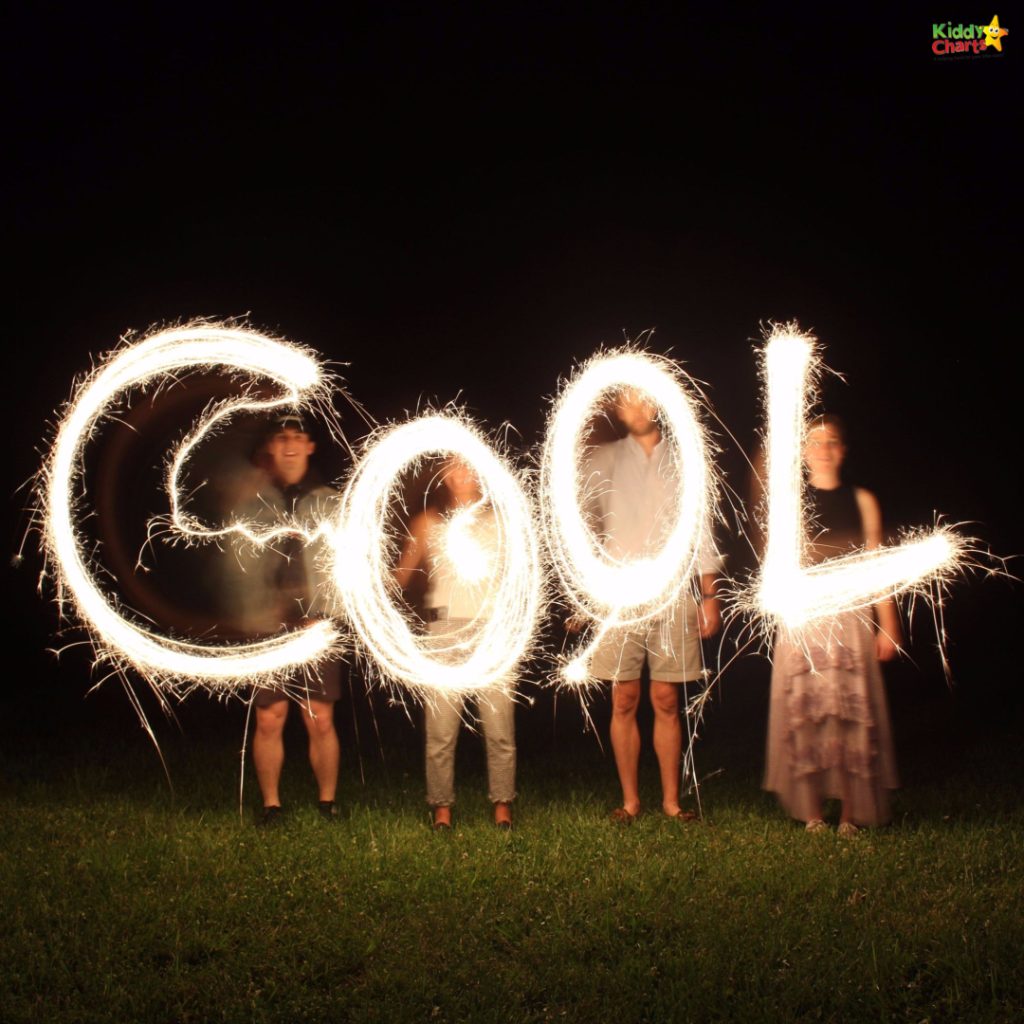 Four people stand on grass, waving sparklers to spell "COOL" in glowing light against a dark background.
