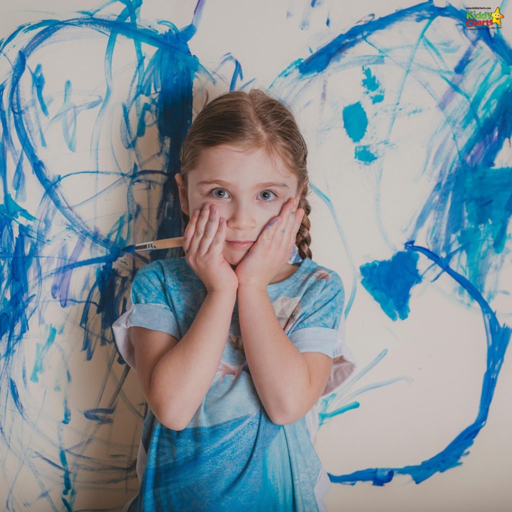 A child in a blue shirt stands with hands on cheeks, in front of a wall with blue crayon scribbles.