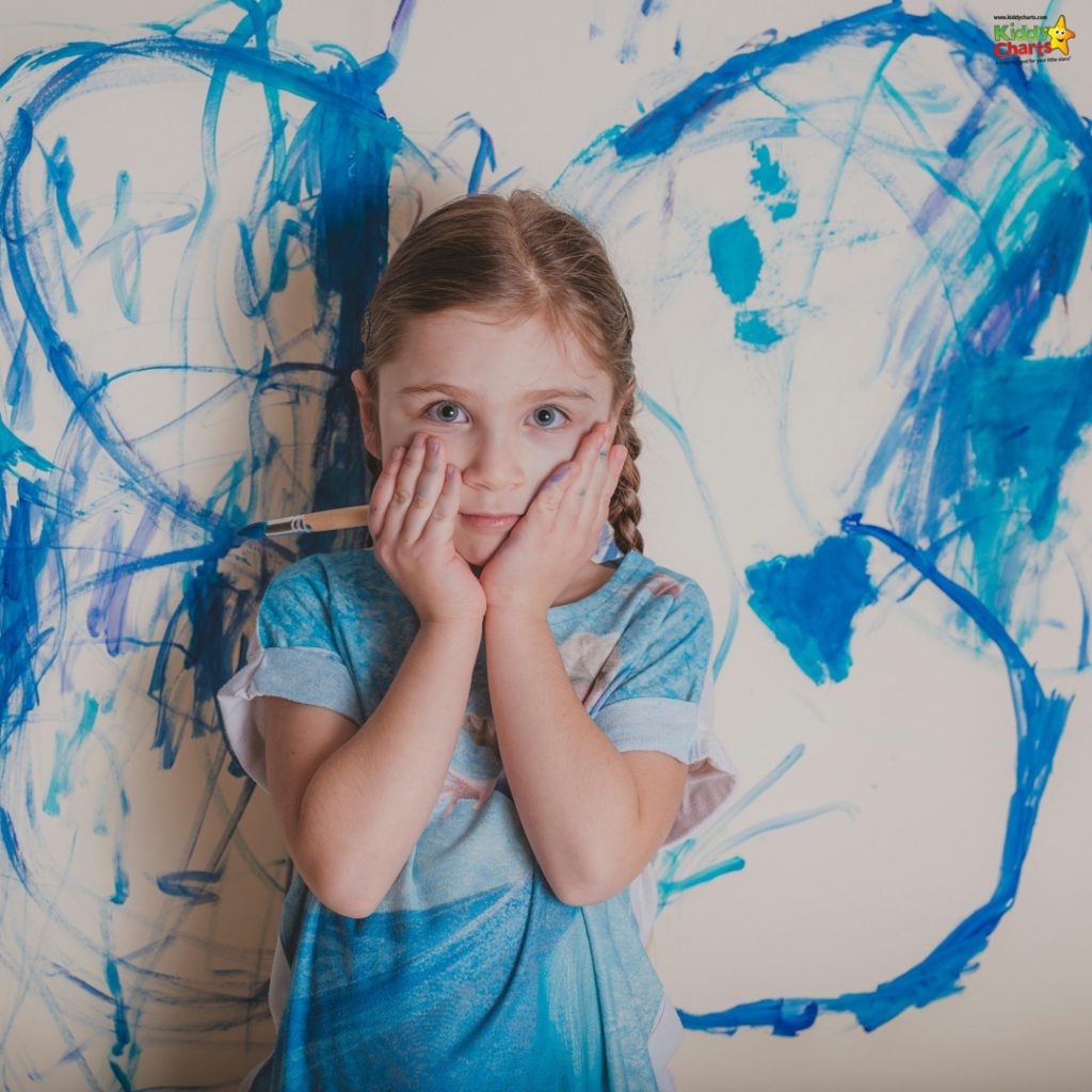 A child with hands on cheeks stands before a wall covered in blue crayon scribbles, wearing a colorful shirt, looking surprised.