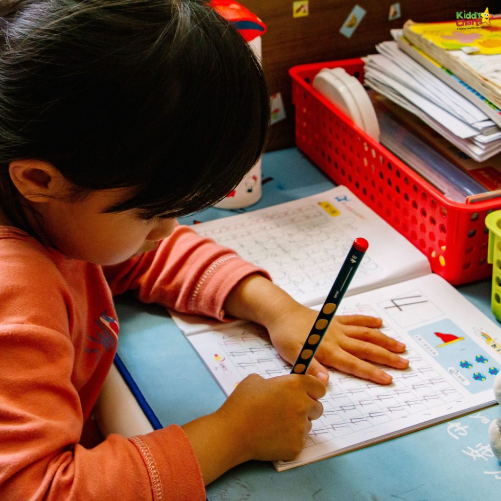 A child is practicing writing with a pencil on a blue desk, surrounded by study materials and a red plastic organizer.