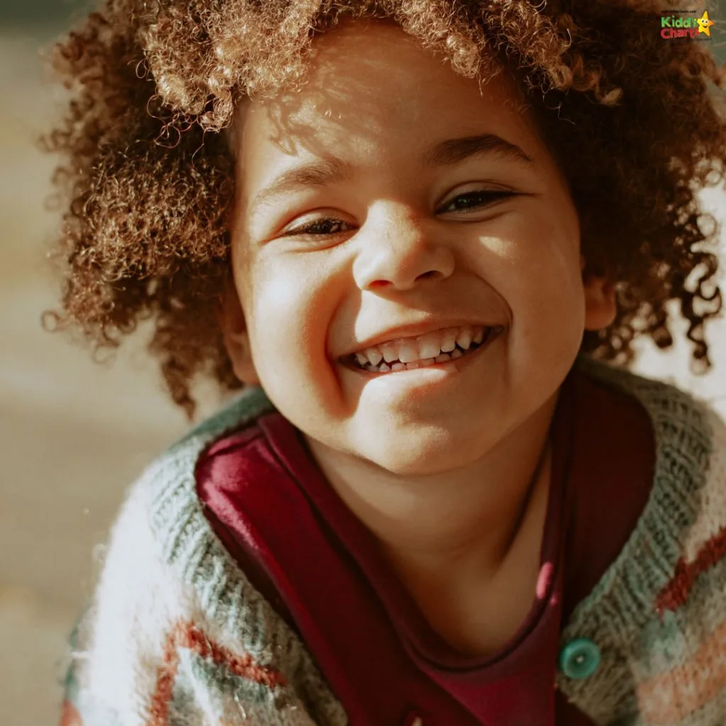 Smiling child with curly hair and cozy sweater in warm sunlight. Joyful expression fills the image with happiness and charm, creating a lively atmosphere.