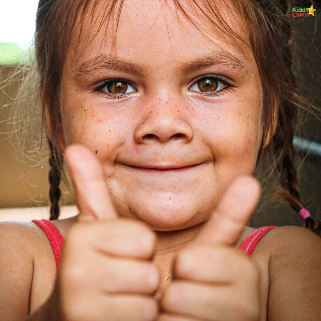 A child with braided hair smiles, giving two thumbs up, wearing a pink top. The background is blurred and features a logo.