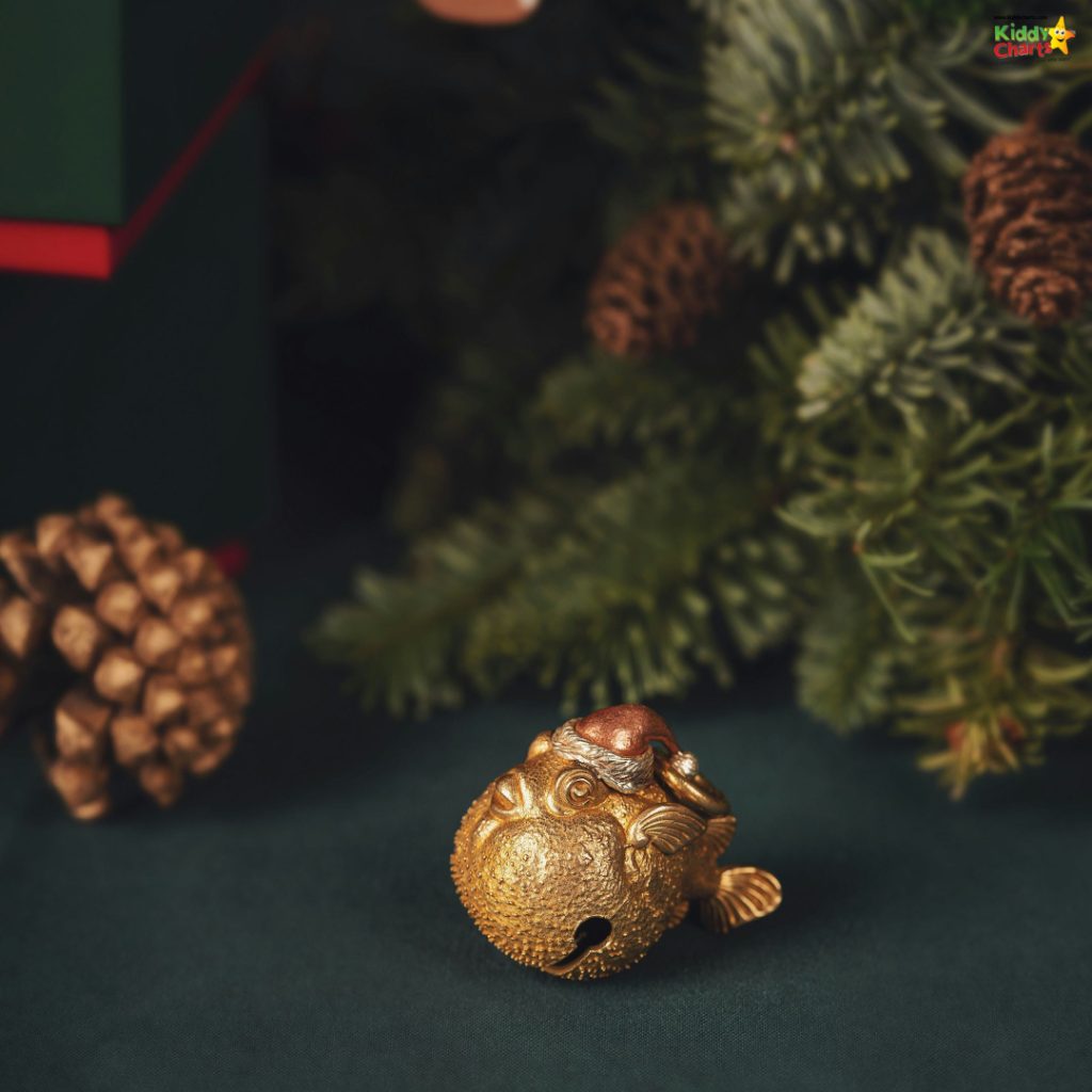 A golden pufferfish ornament with a Santa hat, surrounded by pinecones and evergreen branches on a dark green surface, evokes a festive atmosphere.